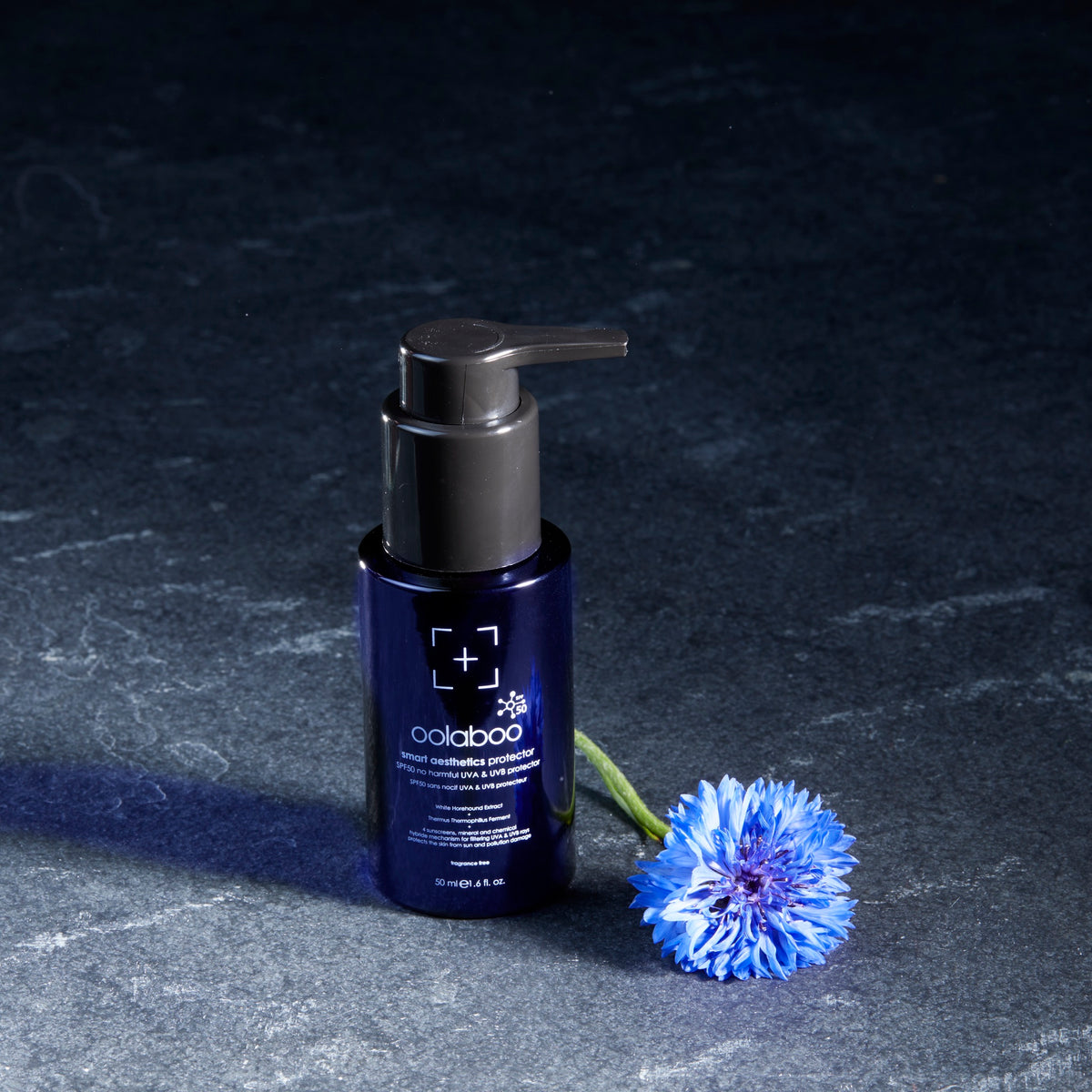 Oolaboo Smart Aesthetics SPF50 Protector frasco sobre pizarra con aciano